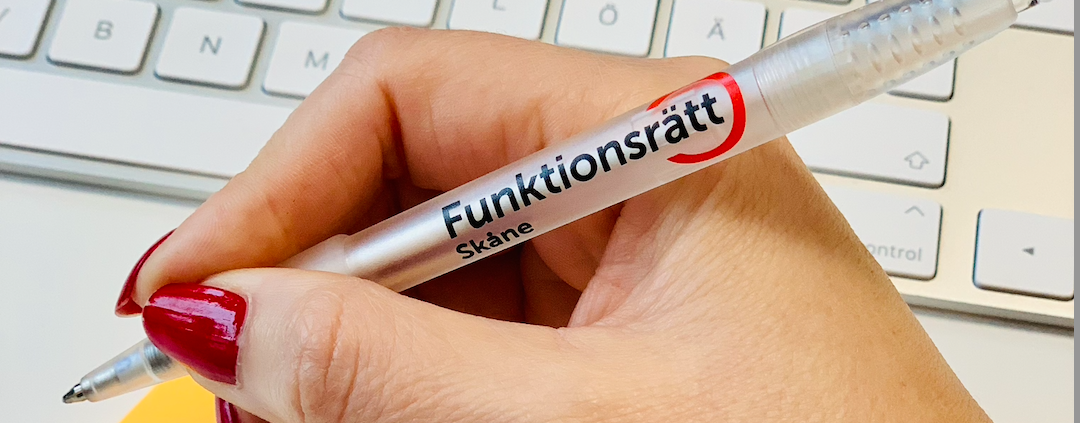 Bild på en hand som håller en penna. På pennan syns Funktionsrätt Skånes logga och bakgrunden skymtas ett tangentbord.