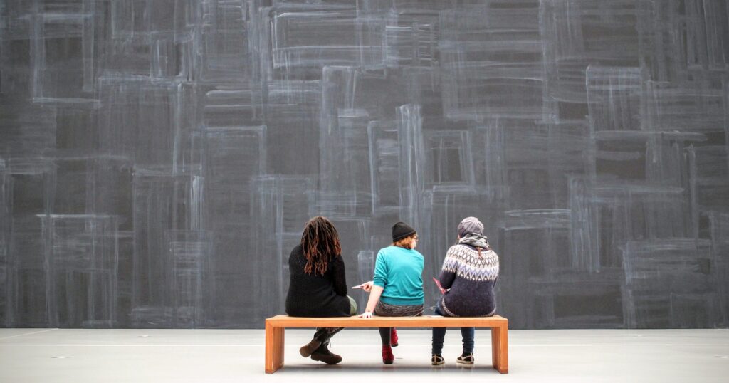 Tre unga människor ses bakifrån då de sitter på en bänk framför stormönstrad vägg.
