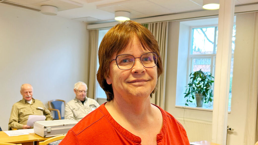 Lena Landin står mitt i ett mötesrum. Bakom syns två män bakom ett bord.