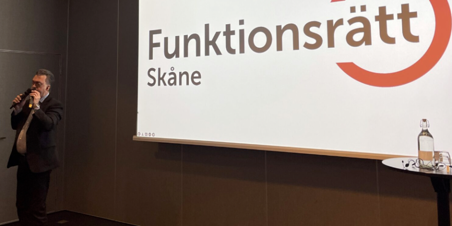 En man med mikrofon står bredvid en stor skärm med Funktionsrätt Skånes logga.